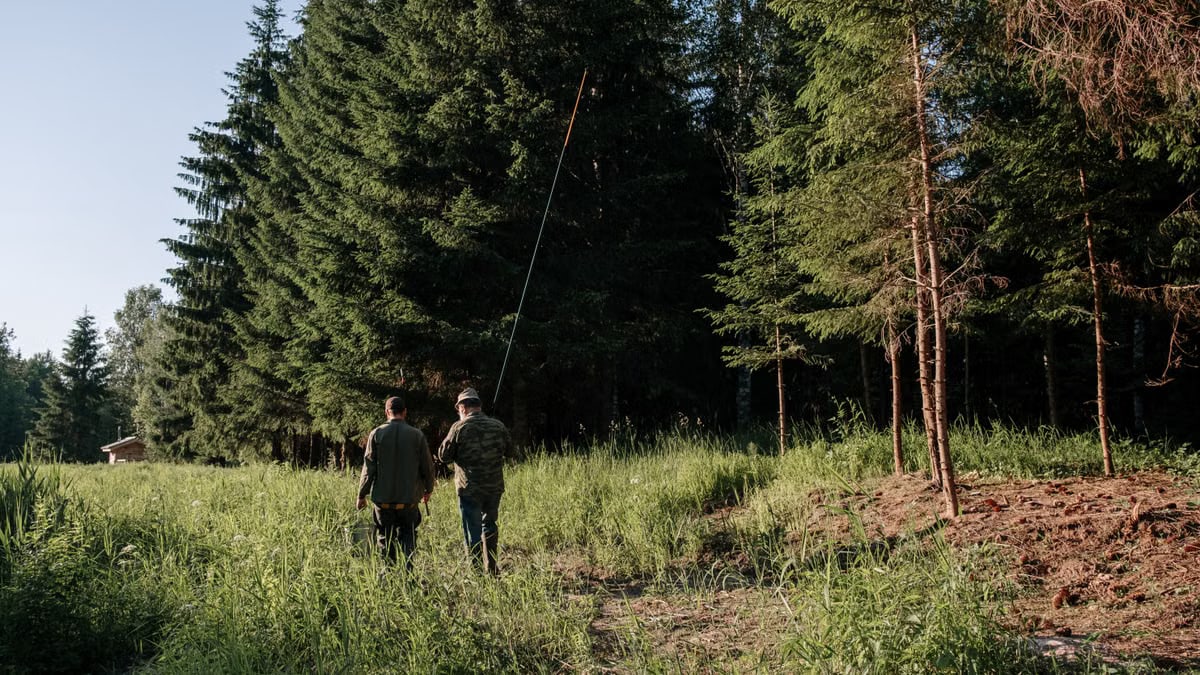 Two men with fishing gear walking through a sunlit forest clearing.